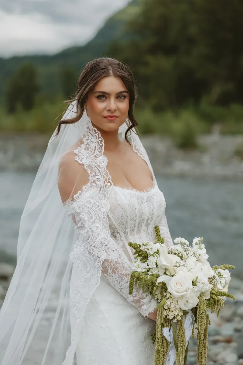 Bride in a lace veil holding her bouquet by the river