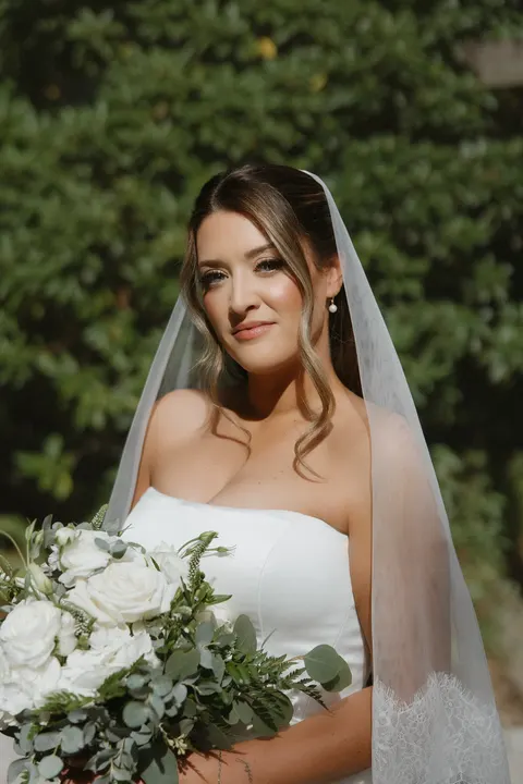 Bride in a sheer veil smiling softly, holding white roses